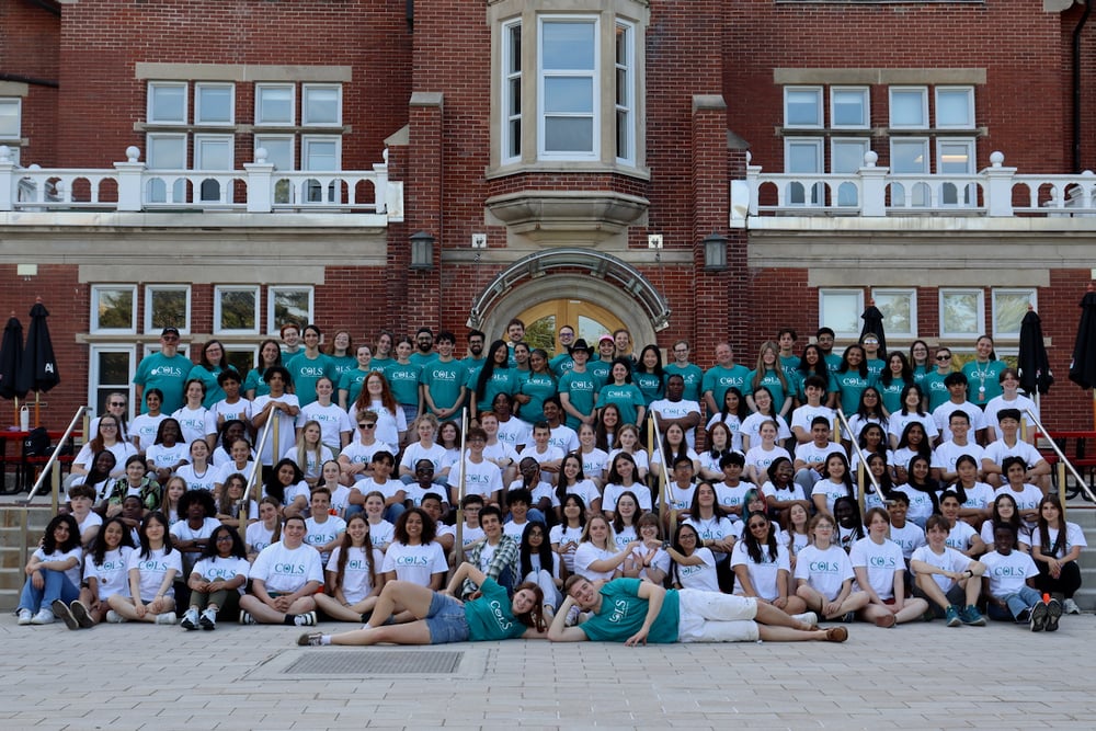 The whole COLS group, in white and teal T-shirts, posing together on the wide steps of a red-brick building