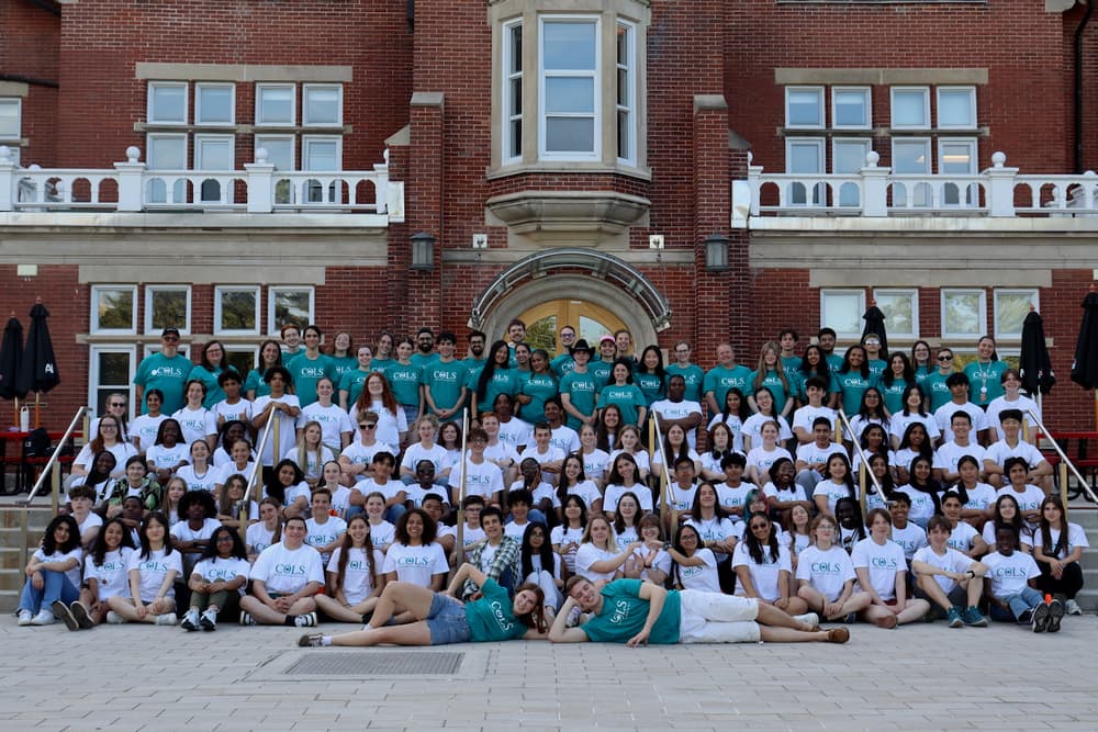 The whole COLS group, in white and teal T-shirts, posing together on the wide steps of a red-brick building