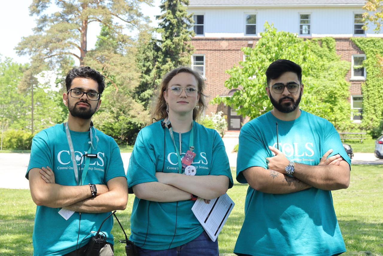 Three COLS staff in teal T-shirts standing on a lawn with their arms crossed, smiling